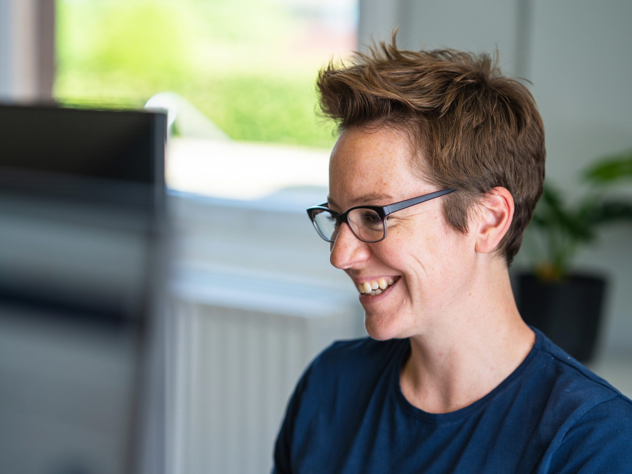 A woman wearing glasses is smiling at a computer while doing content research for a design agency.
